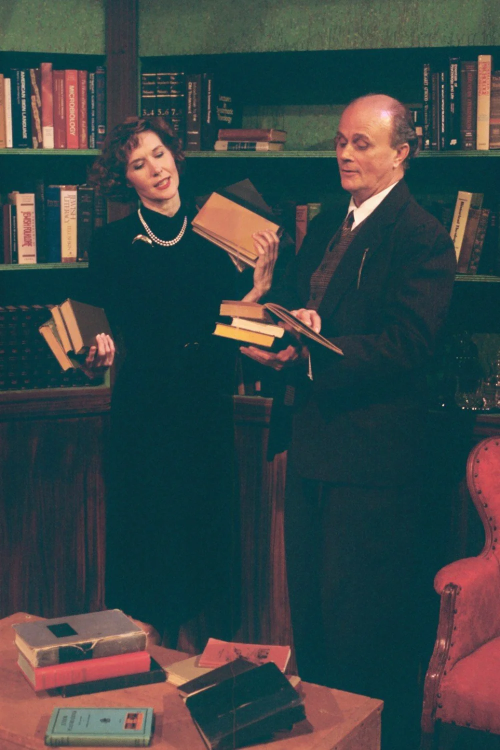 Two people in a library holding books in front of a bookshelf, with a red chair nearby and additional books on a table.
