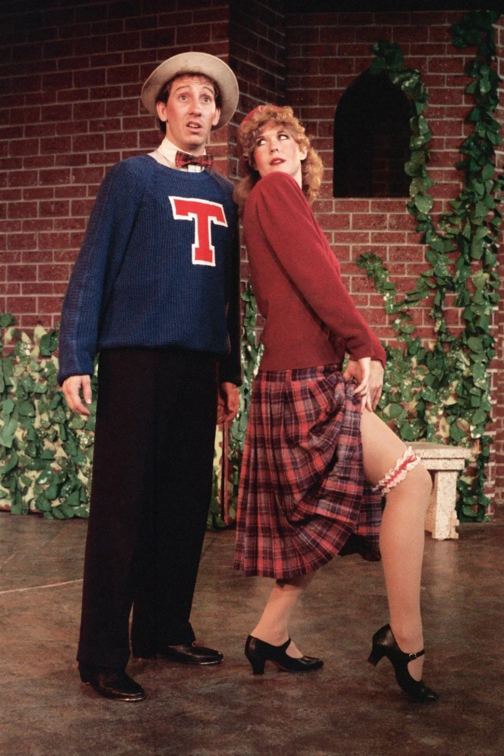 Two actors in a vintage theater setting, one wearing a sweater with a large "T," and the other in a plaid skirt and showing a garter. They stand in front of a brick wall with ivy.