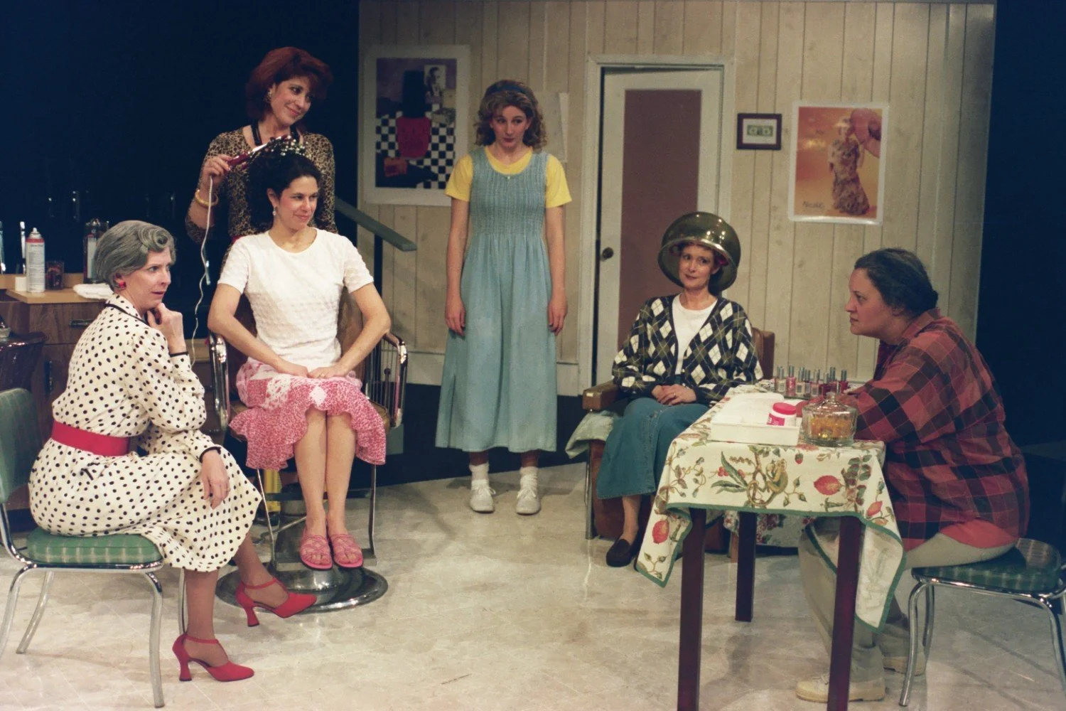 Group of six women in a hair salon setting; one stylist working, others seated, vintage style.