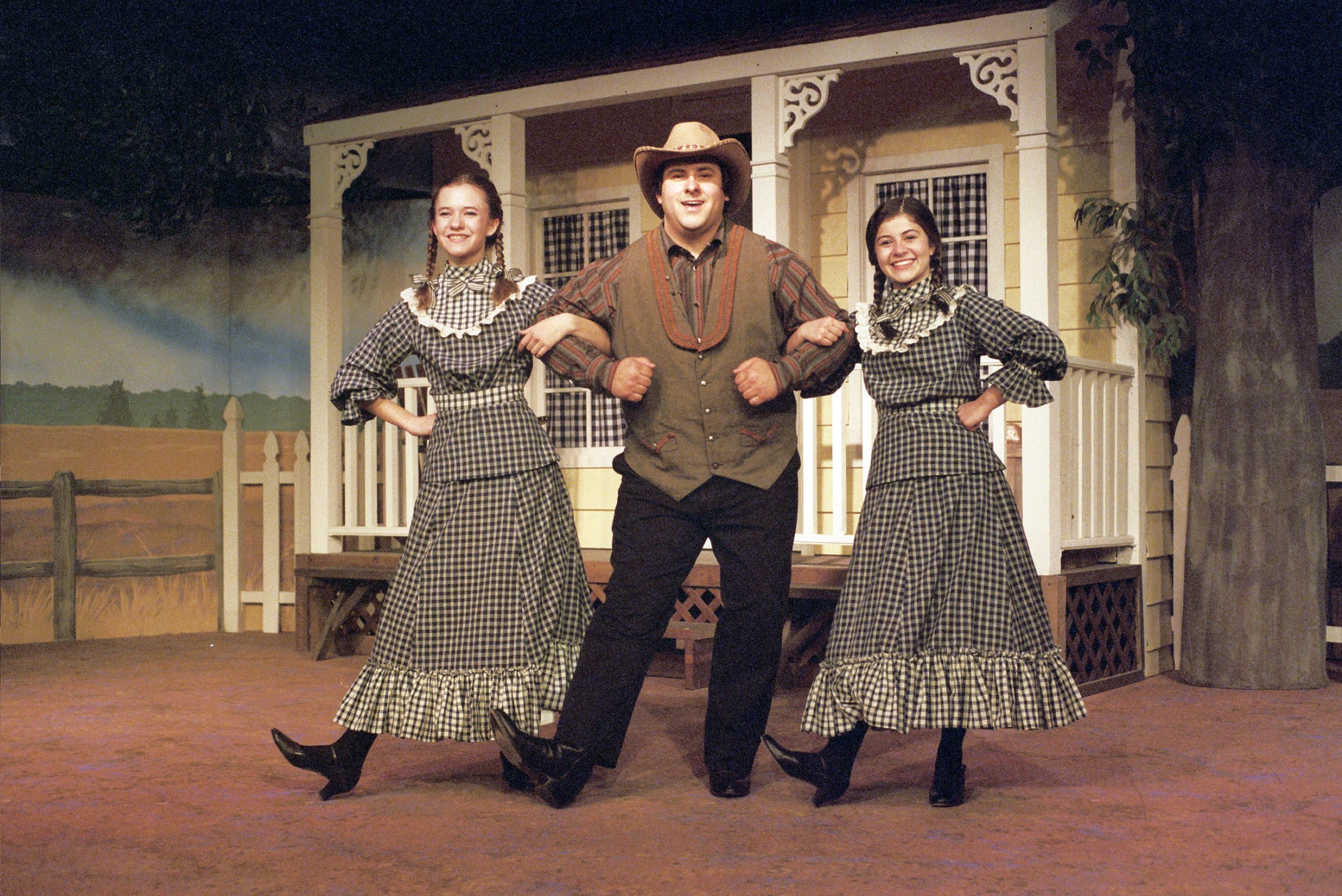 Three actors in Western-themed costumes on stage, with two women in long plaid dresses and a man in a cowboy hat and vest, posing arm-in-arm in front of a house set.