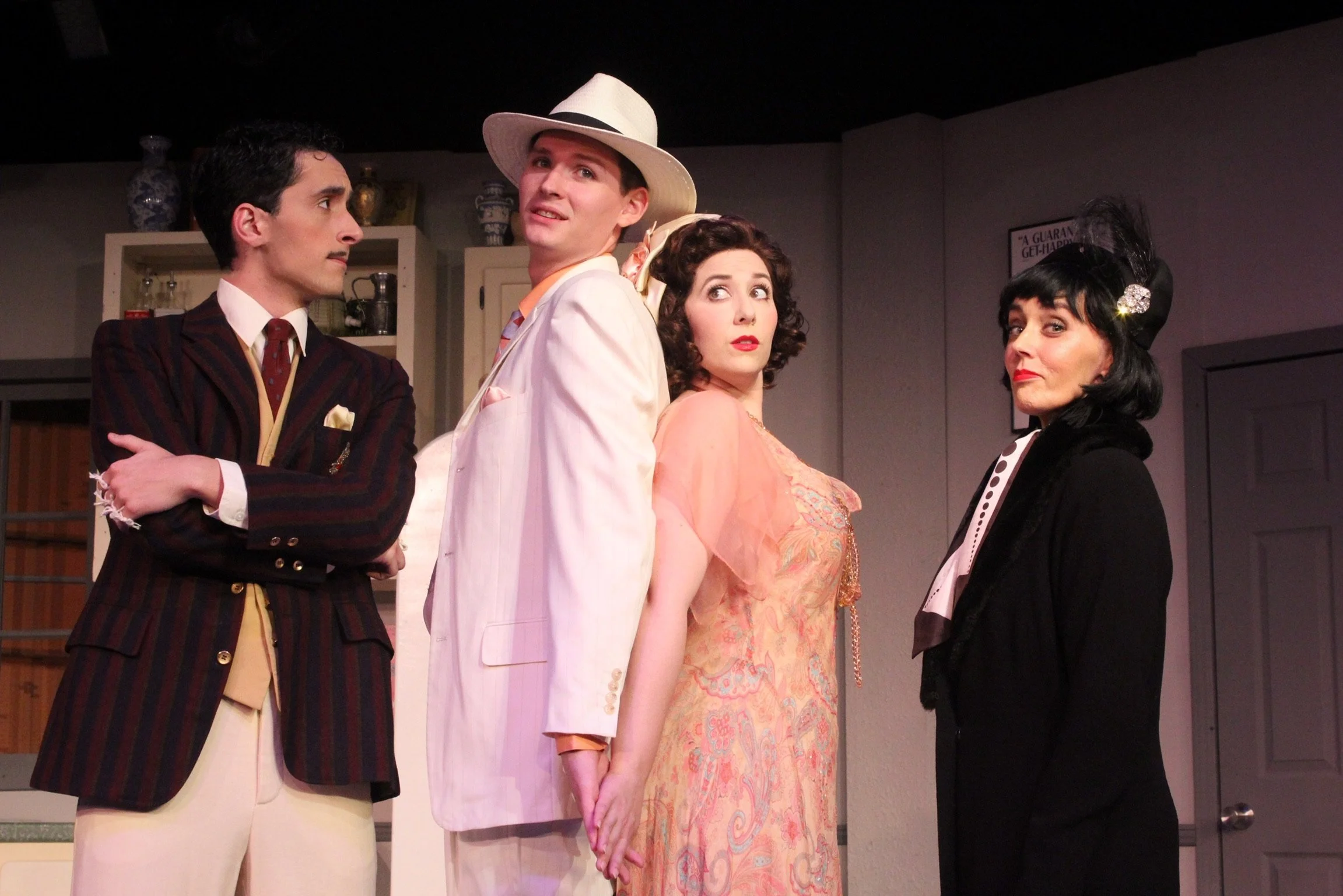 Four actors in theatrical costumes on stage, two men in suits and hats, and two women in vintage dresses. They are posed with varying expressions in front of a kitchen set.