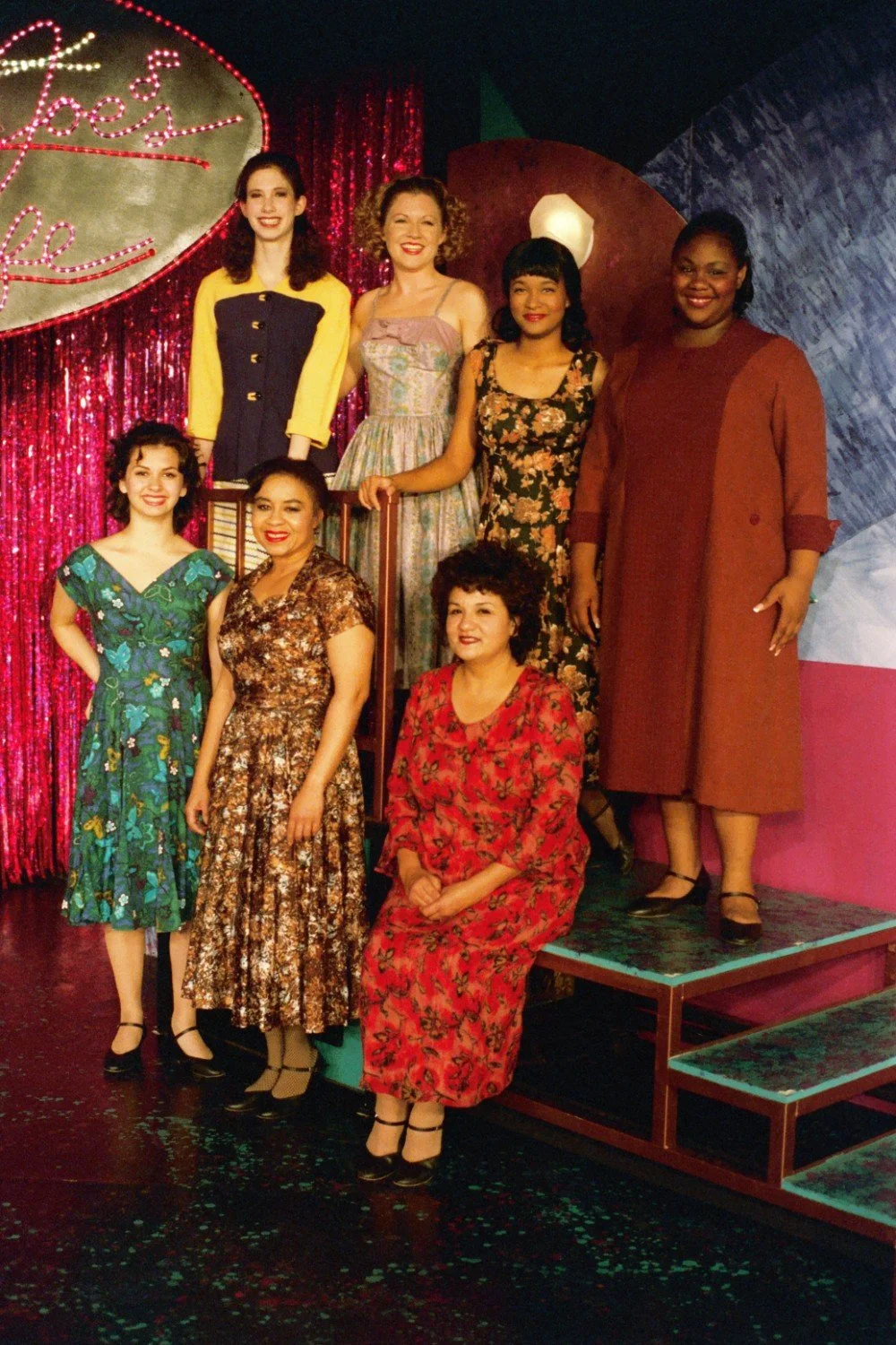 A group of seven women wearing vintage dresses standing and sitting on a colorful stage with a shiny red curtain background and a retro sign.