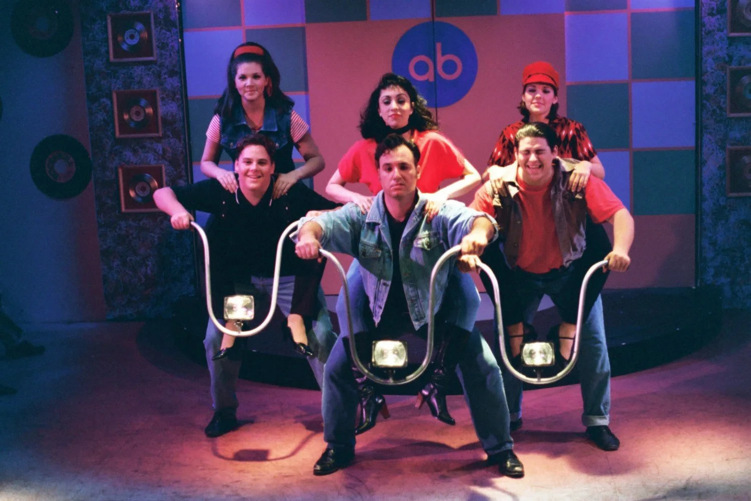 Group of actors performing on set with stage design featuring records and checkerboard pattern backdrop, wearing casual 1950s-style clothing, and posing as if riding motorcycles.