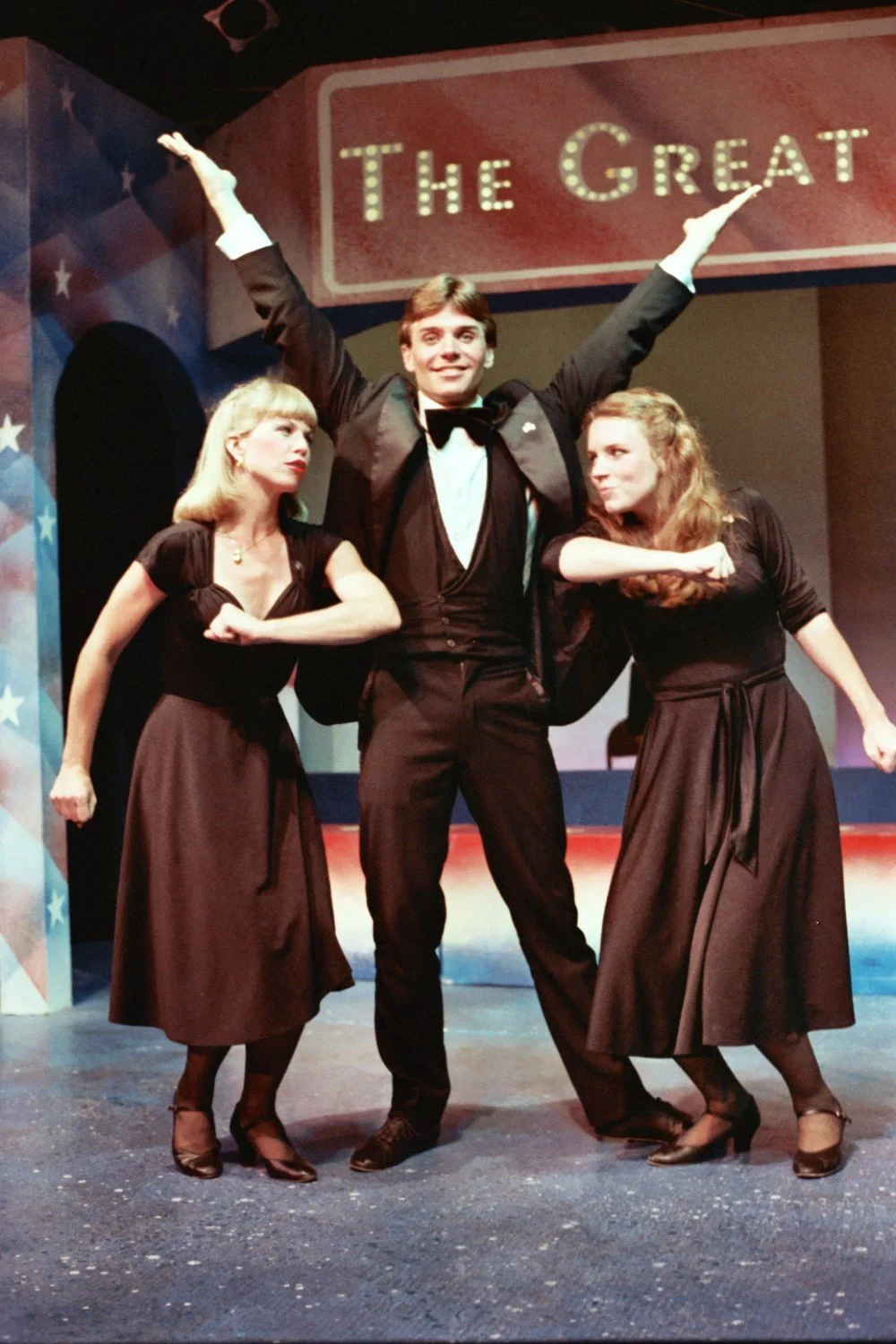Three performers in a theater pose on stage. The man in the center wears a tuxedo, and the two women on either side wear black dresses. The backdrop shows the words 'The Great.'