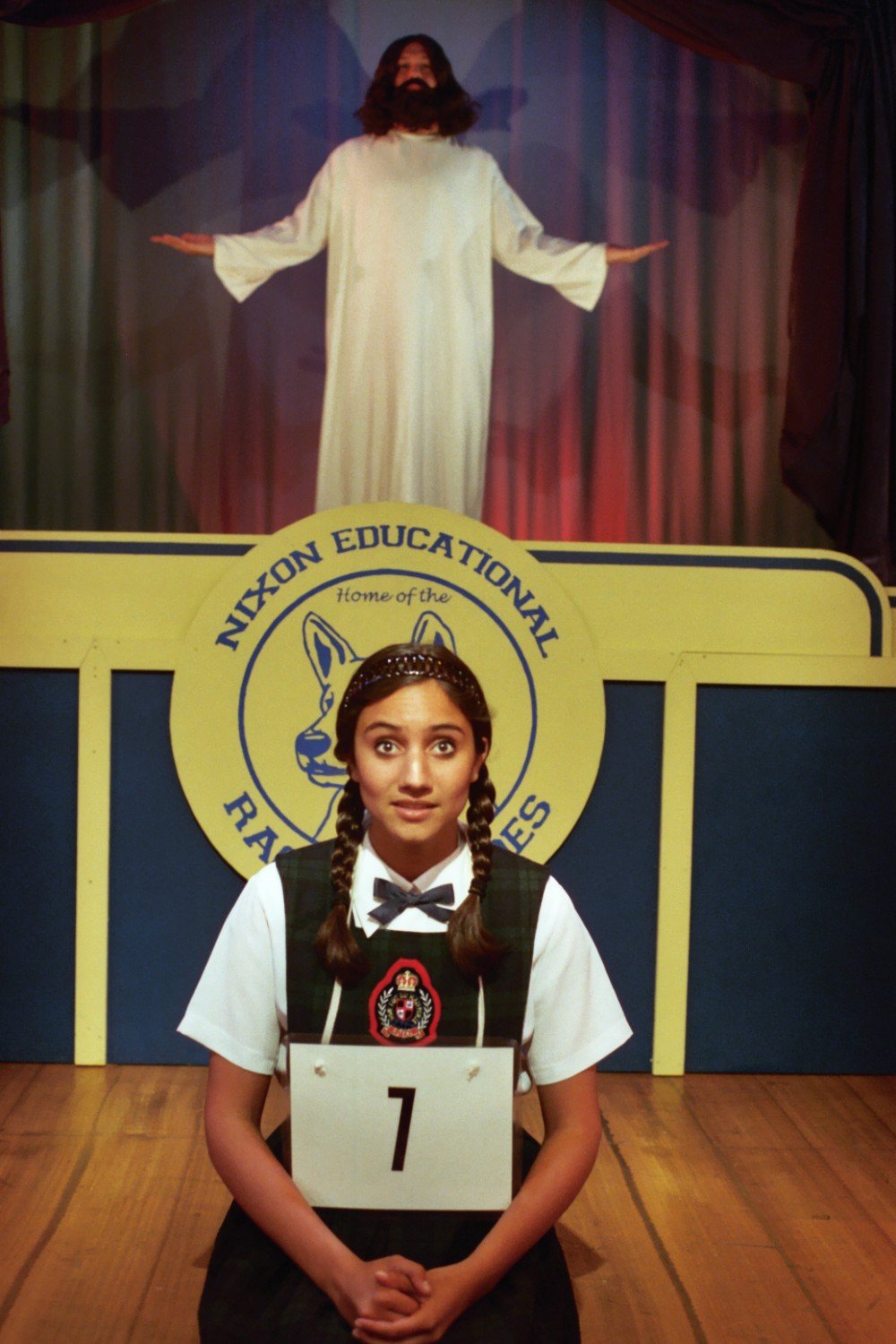 A girl with braided hair and a uniform is kneeling on stage with a number 7 badge, in front of a sign reading "Nixon Educational." Behind her is a person in a white robe with arms outstretched.