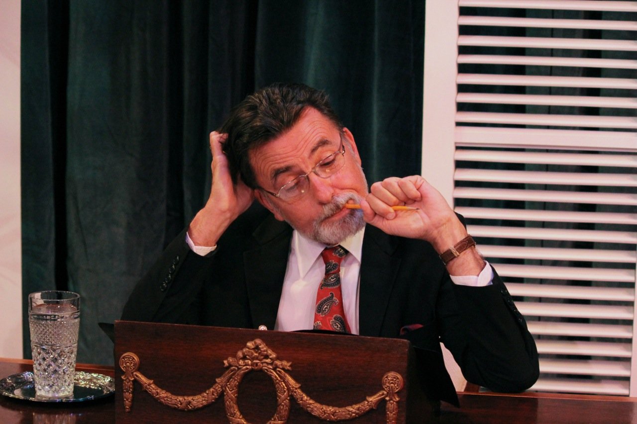 Man in suit sitting at desk with pencil in mouth, hand on head, glass of water, ornate desk