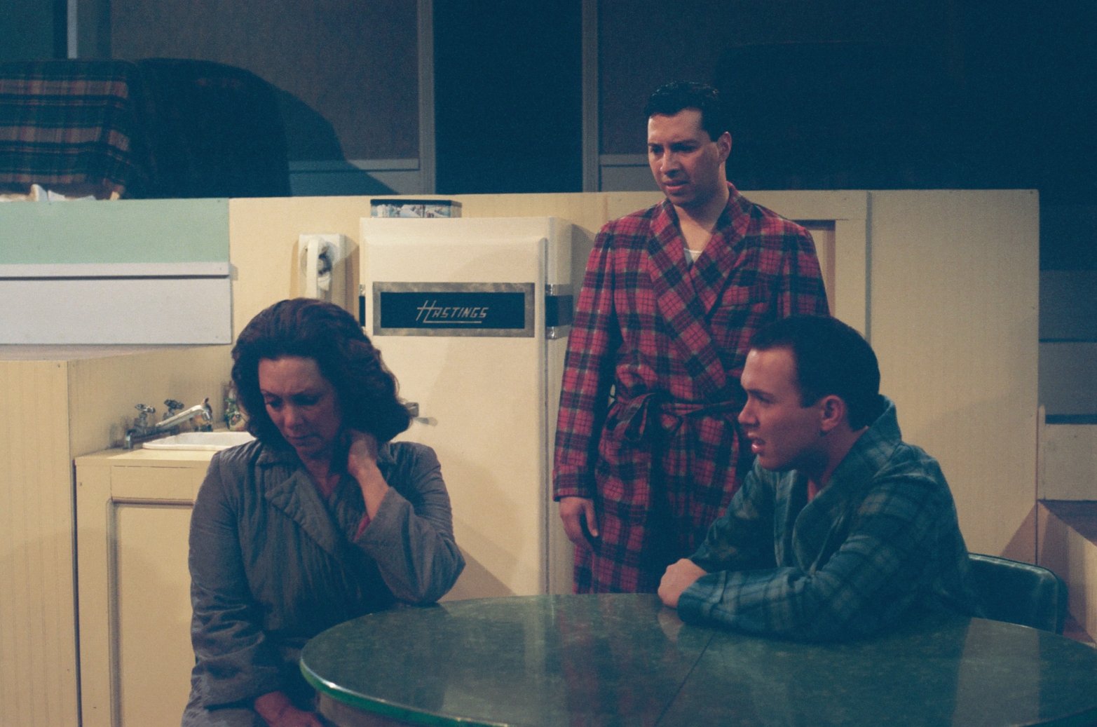 Three people in a dimly lit room, two wearing plaid robes, one sitting and the others standing, appearing in a kitchen-like setting with a vintage fridge and a sink in the background.