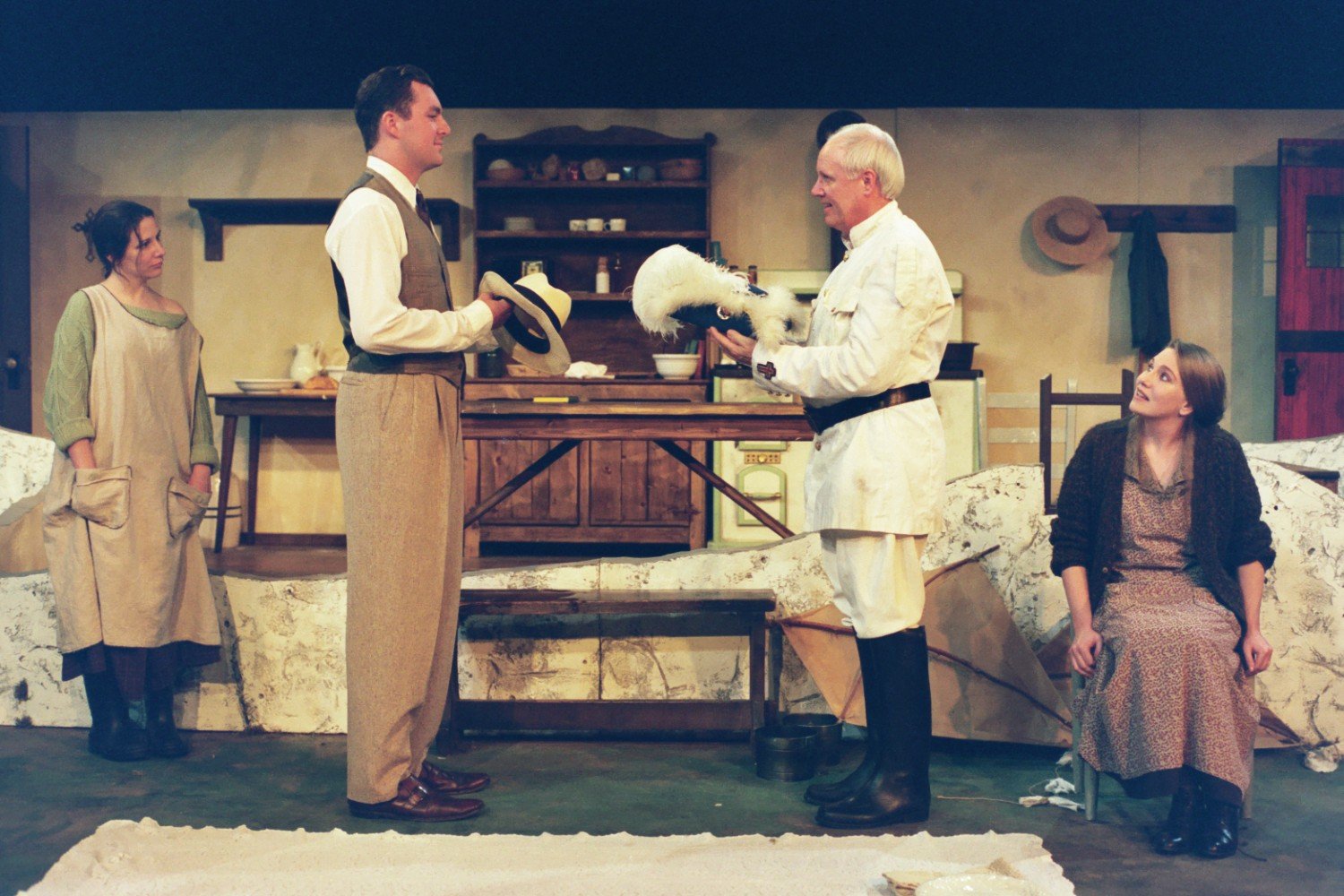 Scene from a theater play with four actors in period costumes in a rustic setting, including a man in a suit holding a hat, a woman in a simple dress looking on, another woman sitting, and an older man dressed in white holding a trophy-like object.