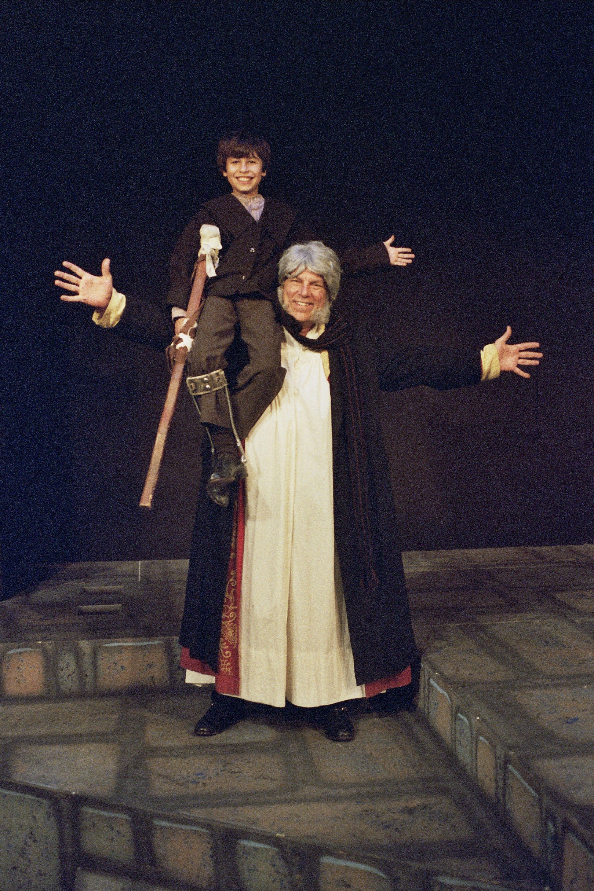 Two people in theatrical costumes, a child dressed as Tiny Tim with a crutch sitting on the shoulders of an older adult dressed as Ebenezer Scrooge, both smiling, against a dark stage background.
