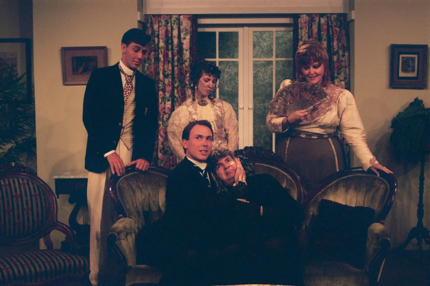A group of people in vintage attire posing in a living room with floral curtains and antique furniture. Two people are seated on a couch, while three others stand behind them. One person is holding a fan.