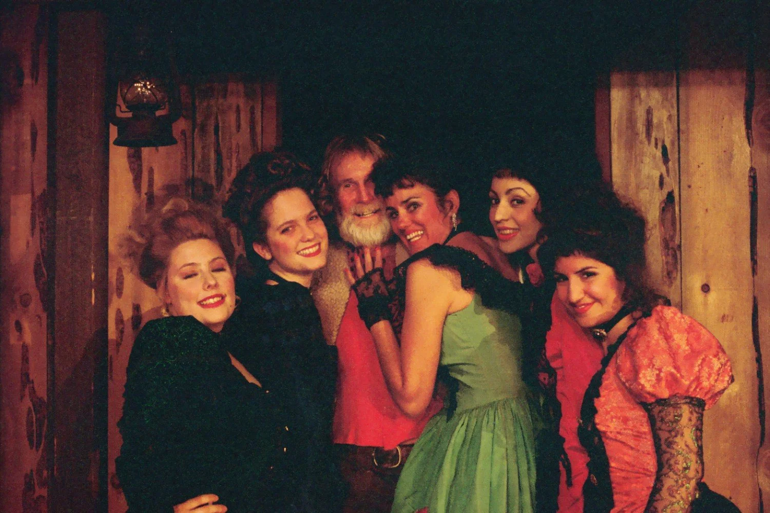 Group of people in vintage-style clothing gathered together indoors, smiling and posing for a photo. The background features wooden walls and a lantern overhead.