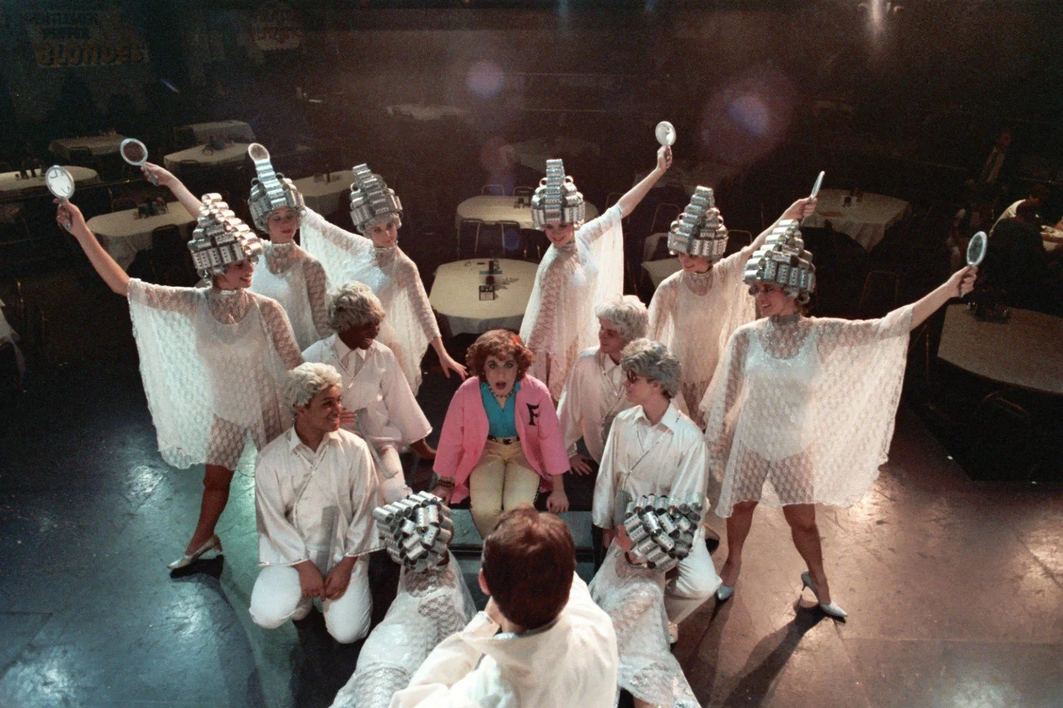 Group of performers on stage, wearing costumes with hair rollers, surrounding a person in a pink jacket, performing a theatrical scene.