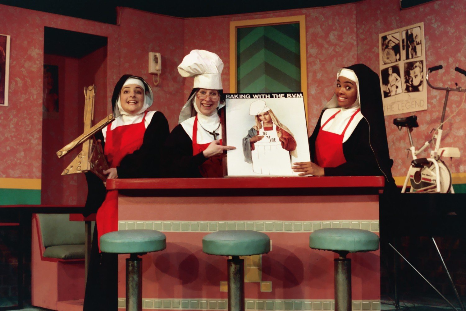 Three women dressed as nuns in a colorful kitchen, holding a poster labeled "Baking with the BVM."