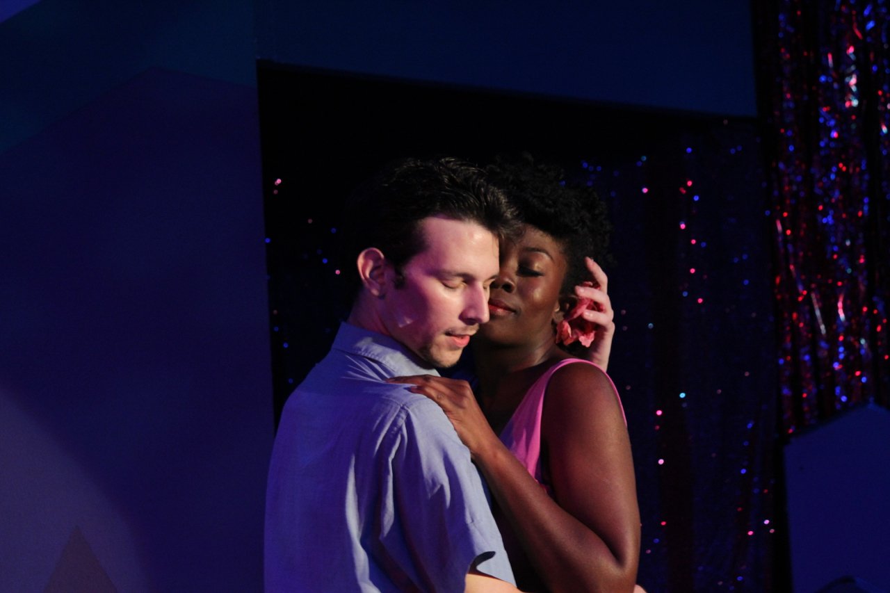 A couple embracing on stage in a dramatic setting, with colorful lighting and a glittery backdrop.