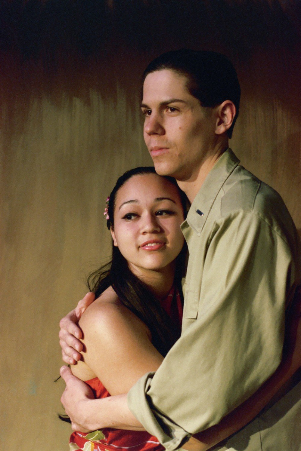 A young couple embracing, with the woman wearing a red patterned dress and the man in a beige shirt, against a plain background.