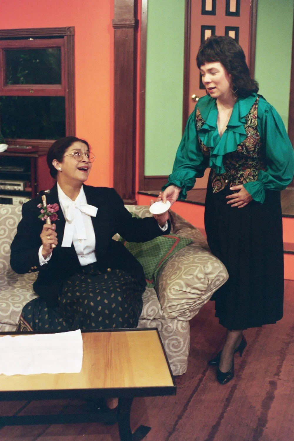 Two women in a living room setting, one sitting on a couch holding a flower, the other standing and offering a cup.