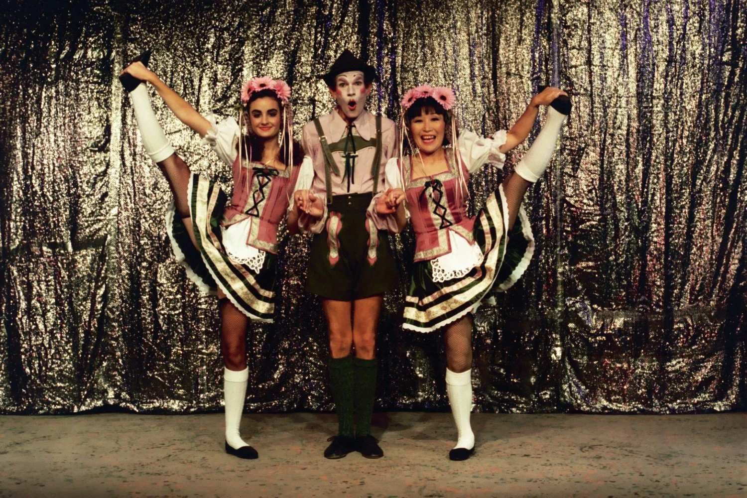 Three performers in costume pose in front of a shimmering curtain, with two women in traditional Bavarian outfits doing high kicks and a clown-like figure in the center.