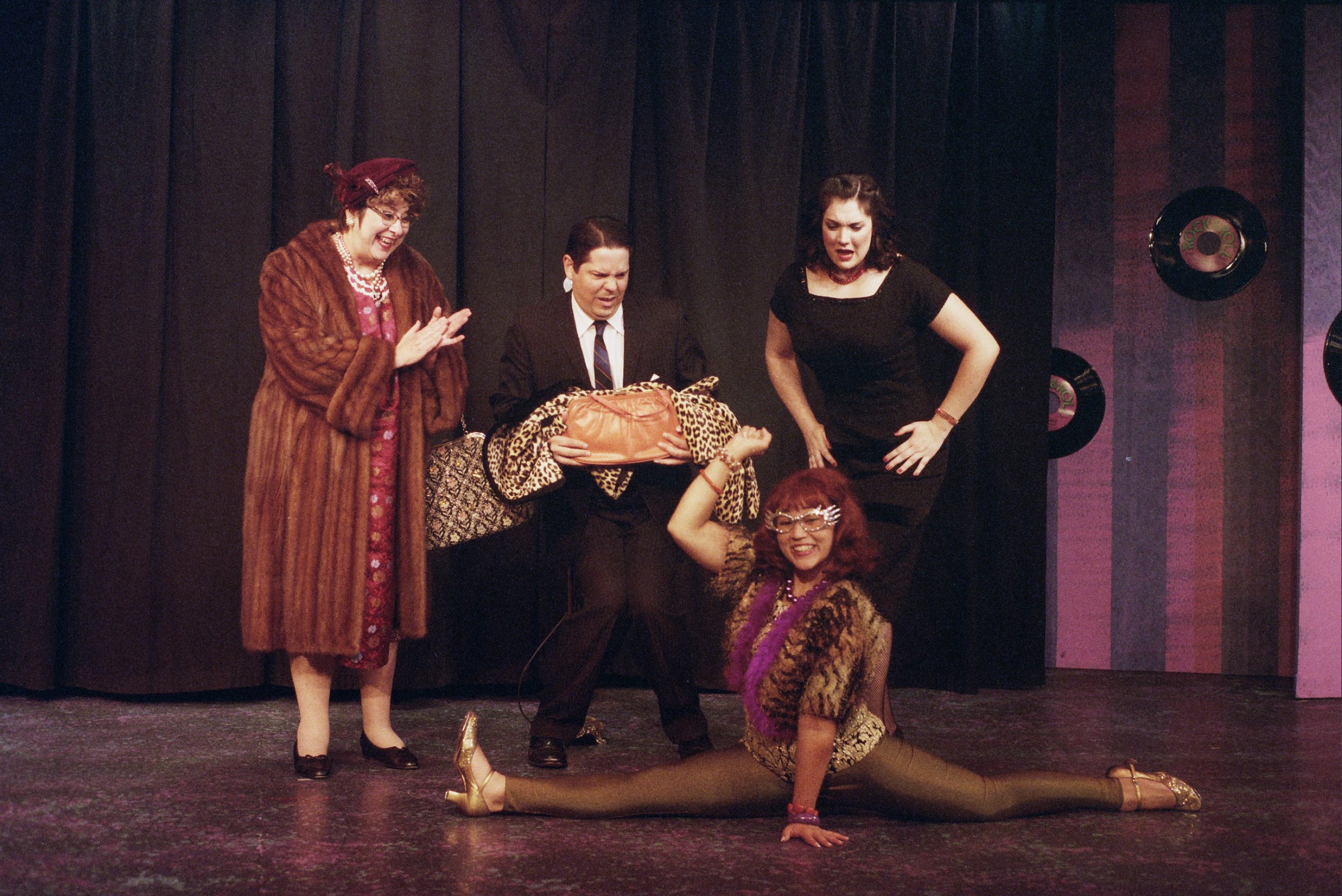 Group of four performers on stage, one woman doing a split in front, others dressed in vintage attire, with a backdrop of curtains and wall decor.