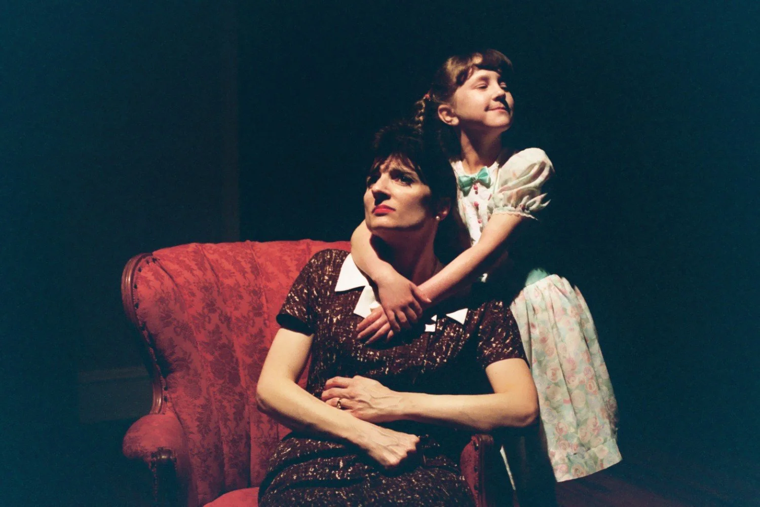 A woman sitting on a vintage red armchair with a young girl standing next to her, embracing her shoulders. The woman is wearing a dark dress with a white collar, and the girl is in a light-colored dress with floral patterns.