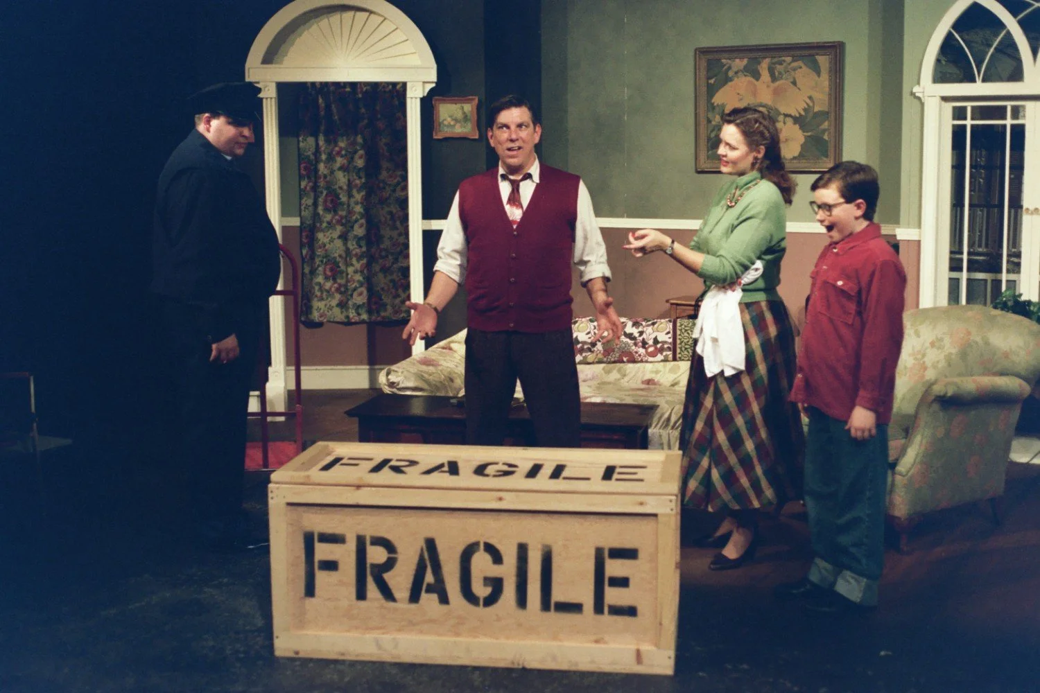 A theatrical scene with four people on stage by a large wooden crate labeled "FRAGILE" in a living room setting. The group includes a man in a police uniform, a man in a red vest, a woman in a green sweater and plaid skirt, and a boy in a red shirt.