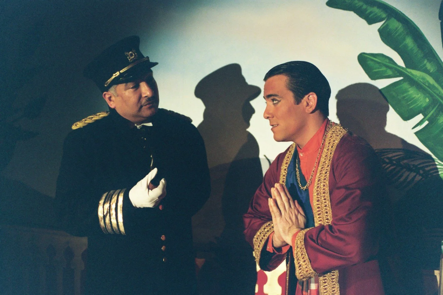 Two men in formal and traditional attire talking on stage, with a shadow of a hat behind them.