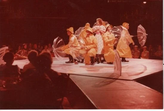 A group of performers in yellow raincoats and hats sit on a stage holding clear umbrellas during a theatrical production, with an audience in the foreground.