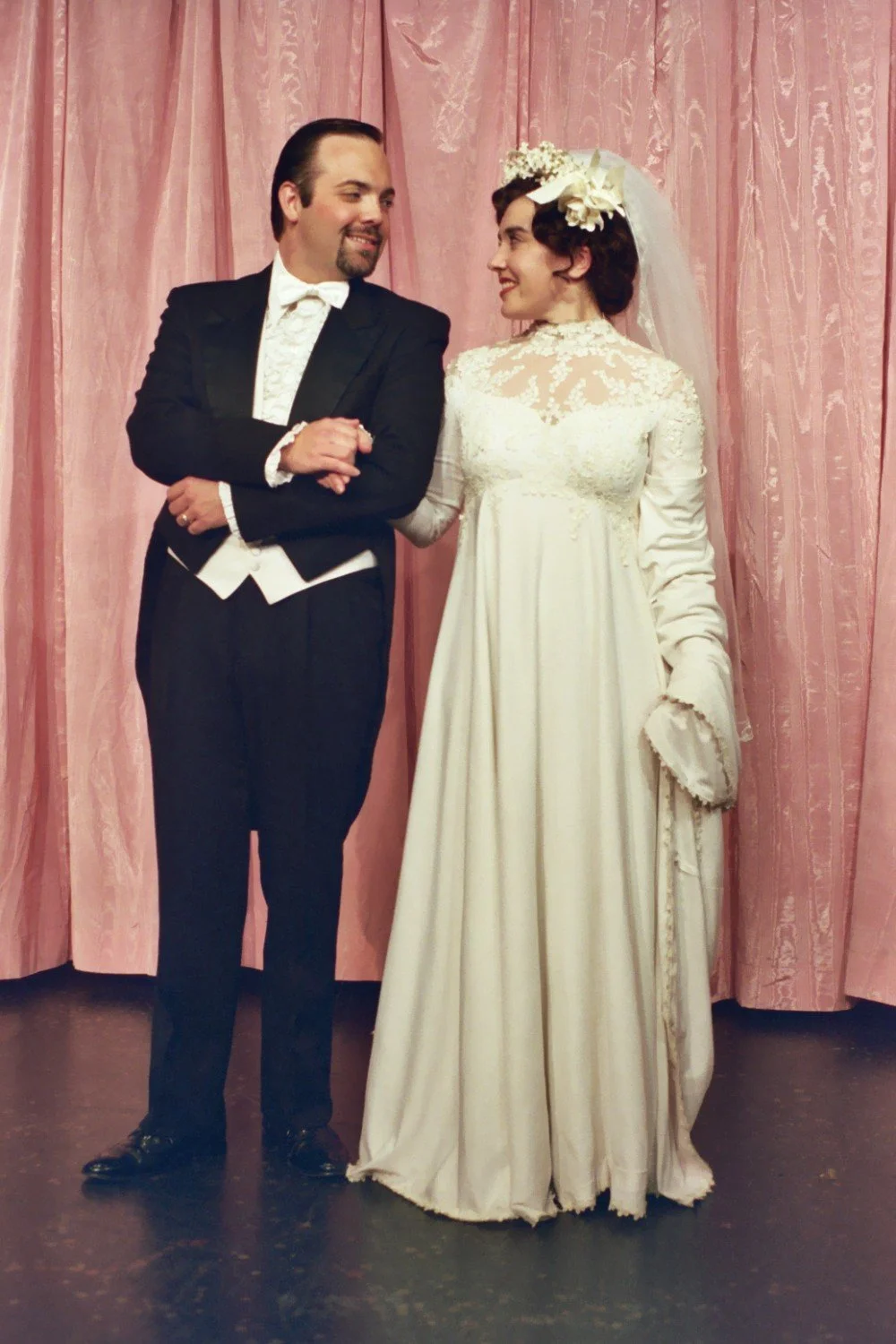 A couple in formal attire; the man is in a tuxedo, and the woman is in a wedding dress with a veil, standing in front of a pink curtain.
