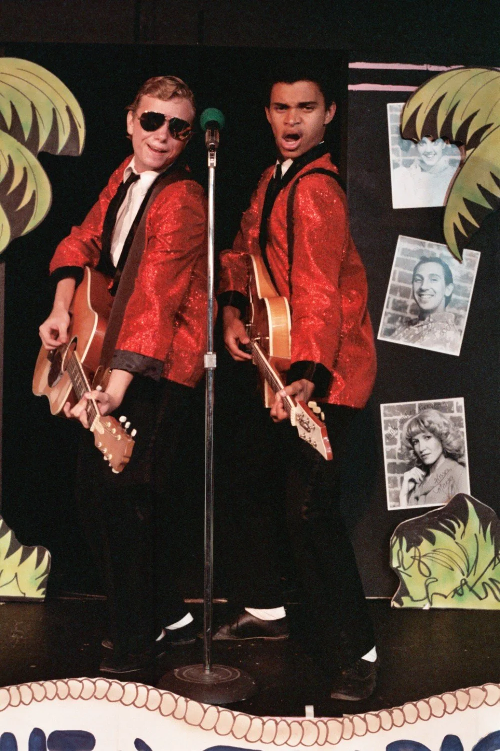 Two performers in red sequined jackets and black pants playing guitars on stage, with palm tree decorations and vintage black-and-white photos in the background.