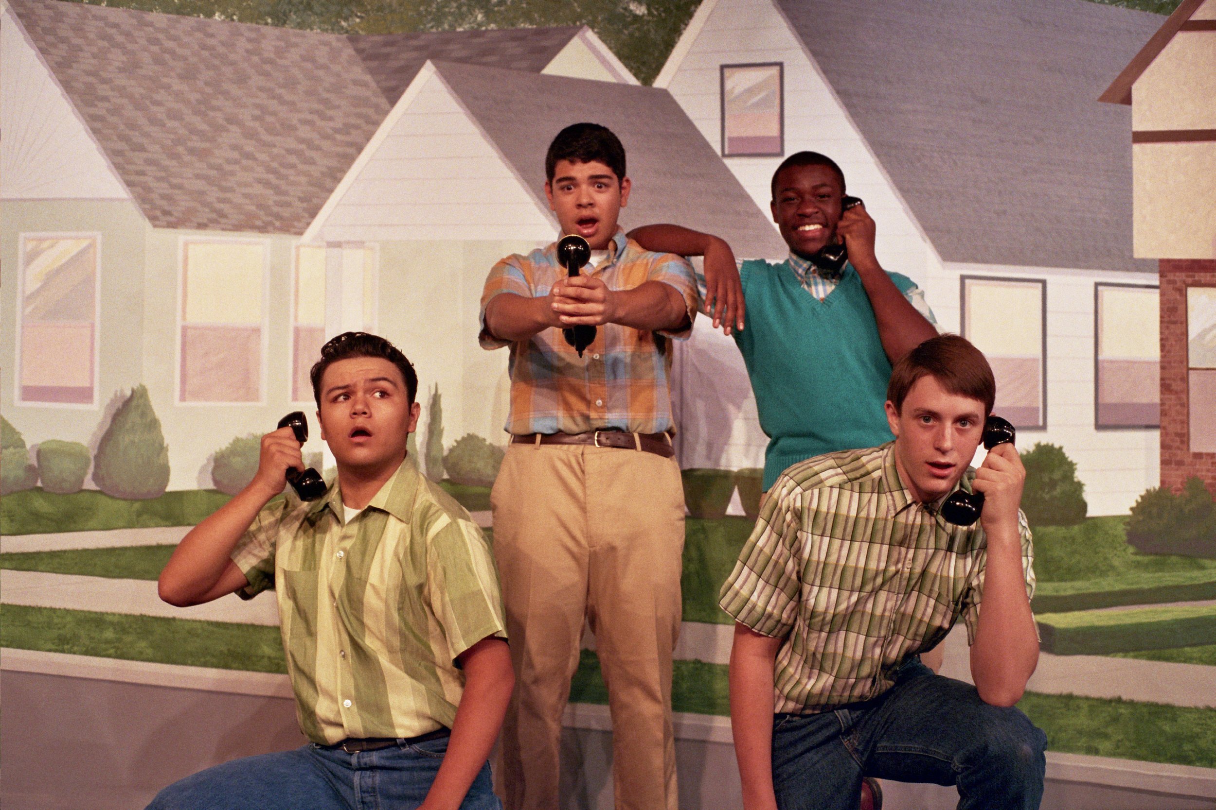 Four young men in vintage-style plaid shirts with telephone receivers, posing humorously in front of a backdrop depicting suburban houses.