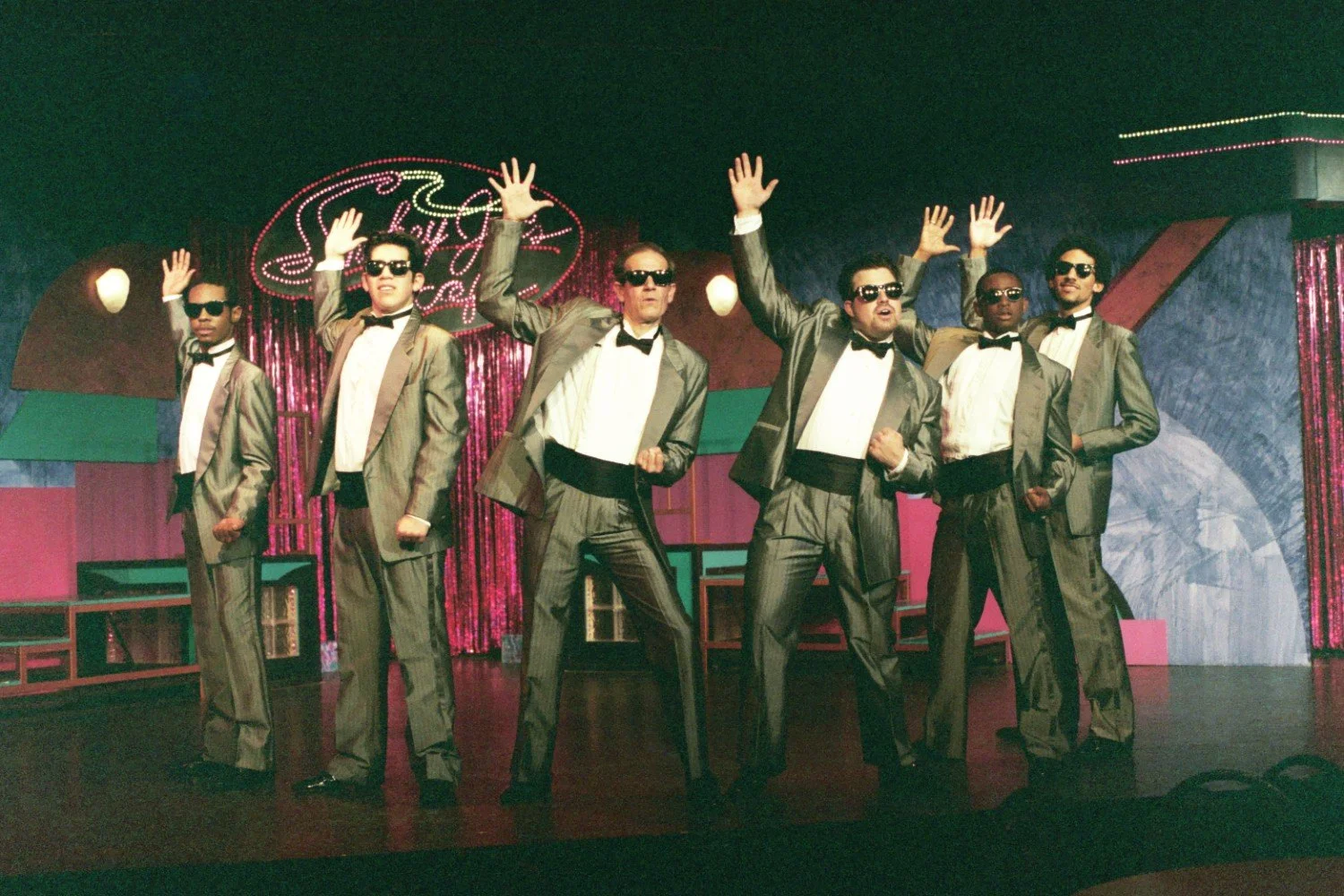 Group of performers in silver suits and sunglasses on stage with a colorful backdrop.