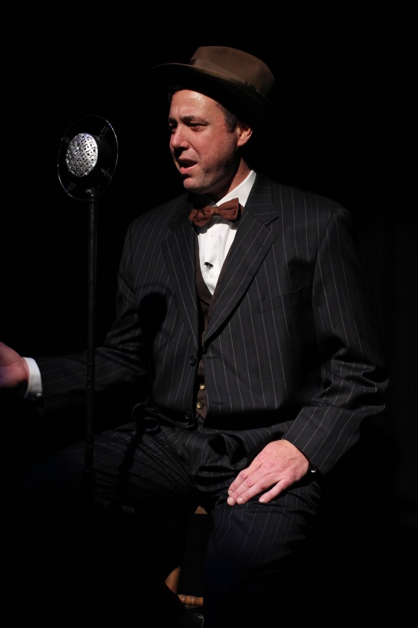 Man in vintage suit and hat speaking into a retro microphone on stage.