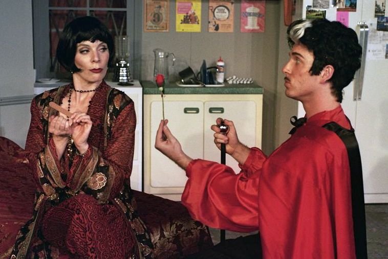 A person in a patterned robe with a bob haircut holds a nail file, while another person in a Dracula costume offers a rose on one knee, in an interior room with a kitchen in the background.