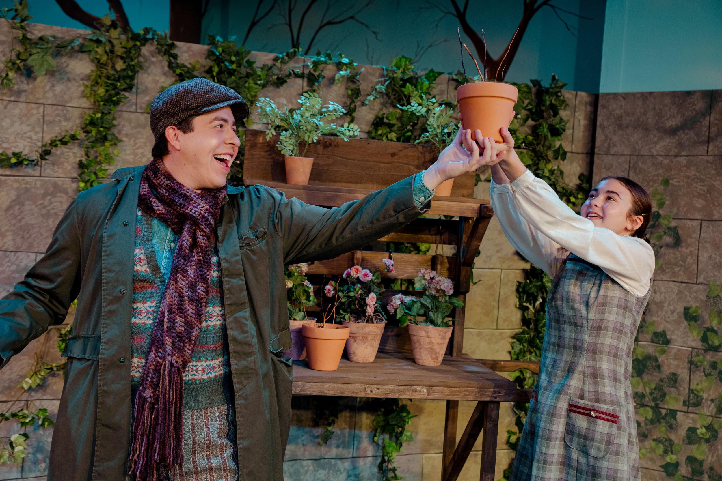 Two people smiling in a garden setting, one holding a plant pot above their head. Background includes a wooden shelf with plants and climbing vines on a stone wall.