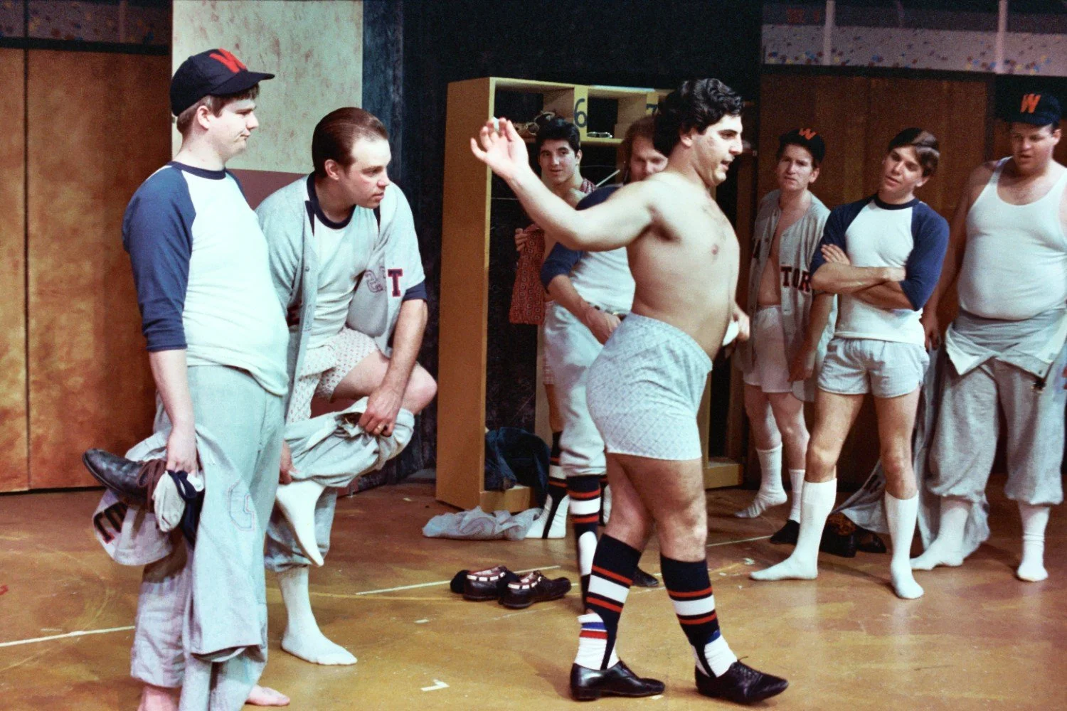 A group of men in a locker room with some wearing athletic uniforms, while others are in undershorts and socks, having a lively conversation.