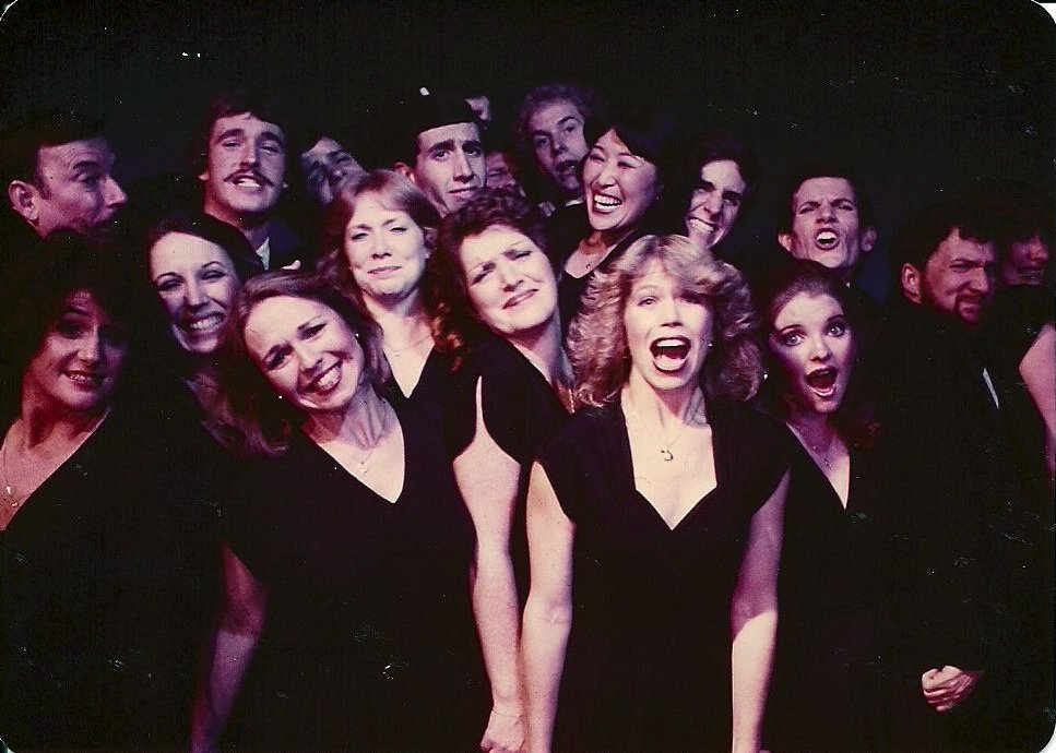 Group of people posing in black outfits against a dark background, smiling and showing various expressions.