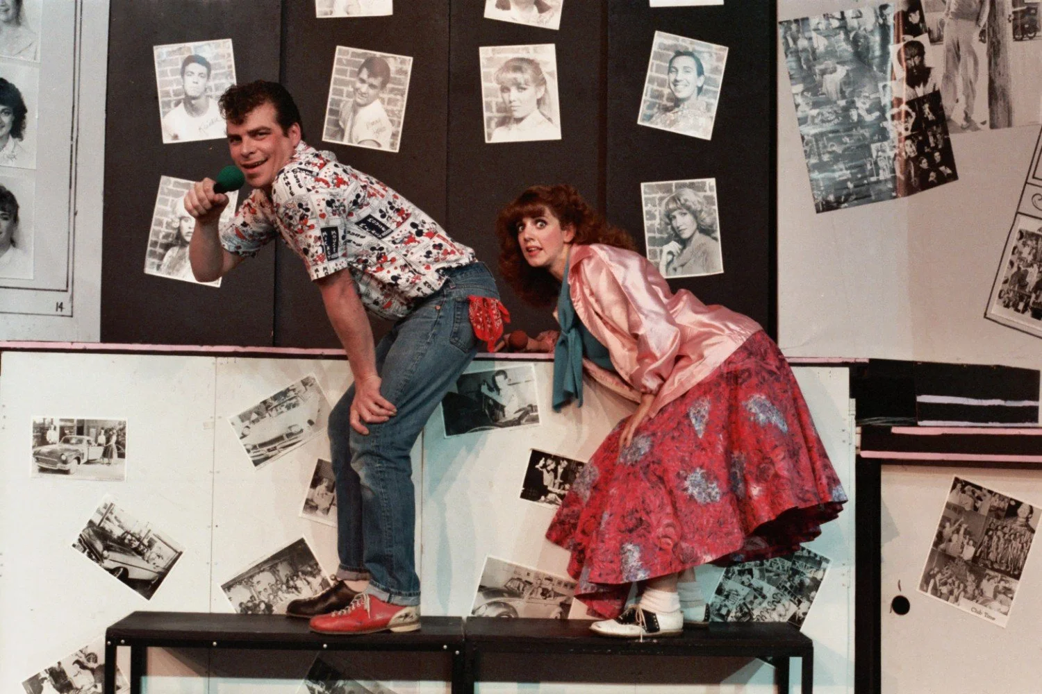 Two actors perform on stage with a backdrop covered in black-and-white photos. The man, holding a green microphone, wears a patterned shirt and blue jeans. The woman, dressed in a pink satin jacket and floral skirt, leans over, watching him closely. 