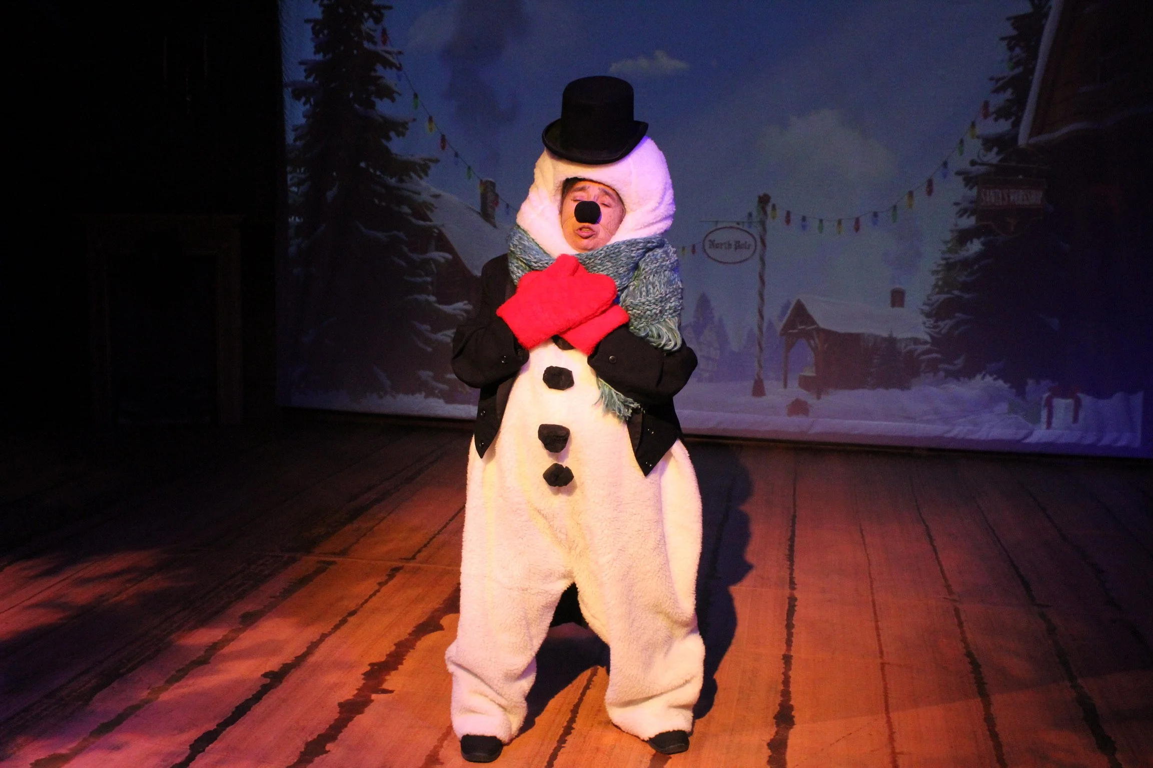Person dressed as a snowman with a top hat, red gloves, and a blue scarf on stage.