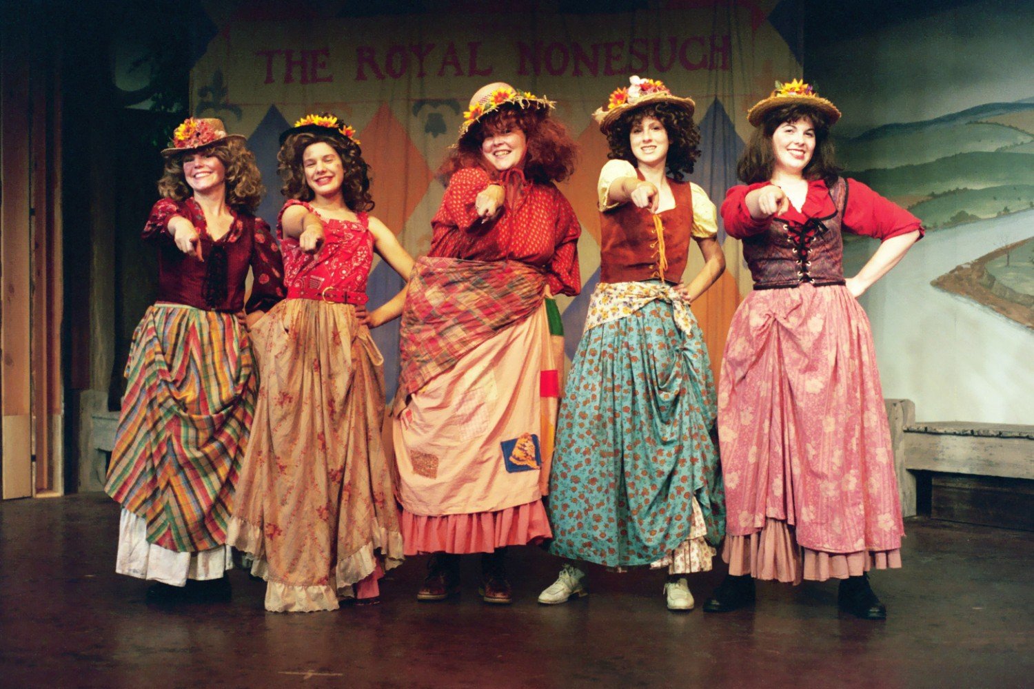 Five women in colorful, vintage-style costumes with hats, standing on stage pointing forward; backdrop reads 'The Royal Nonesuch.'