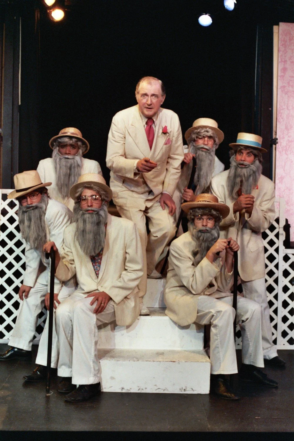 A group of performers in a theatrical setting, wearing light-colored suits, fake gray beards, and hats. They are posed on a multi-level platform against a backdrop with lattice panels, under stage lighting.