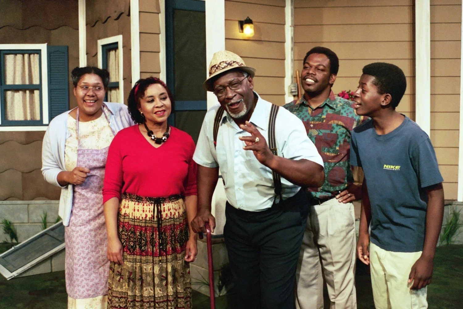 Five people standing in front of a house set, smiling and interacting. The group includes two women, two men, and a boy. One man is wearing a hat and suspenders, holding a cane. They appear engaged and cheerful, as if part of a theatrical scene.