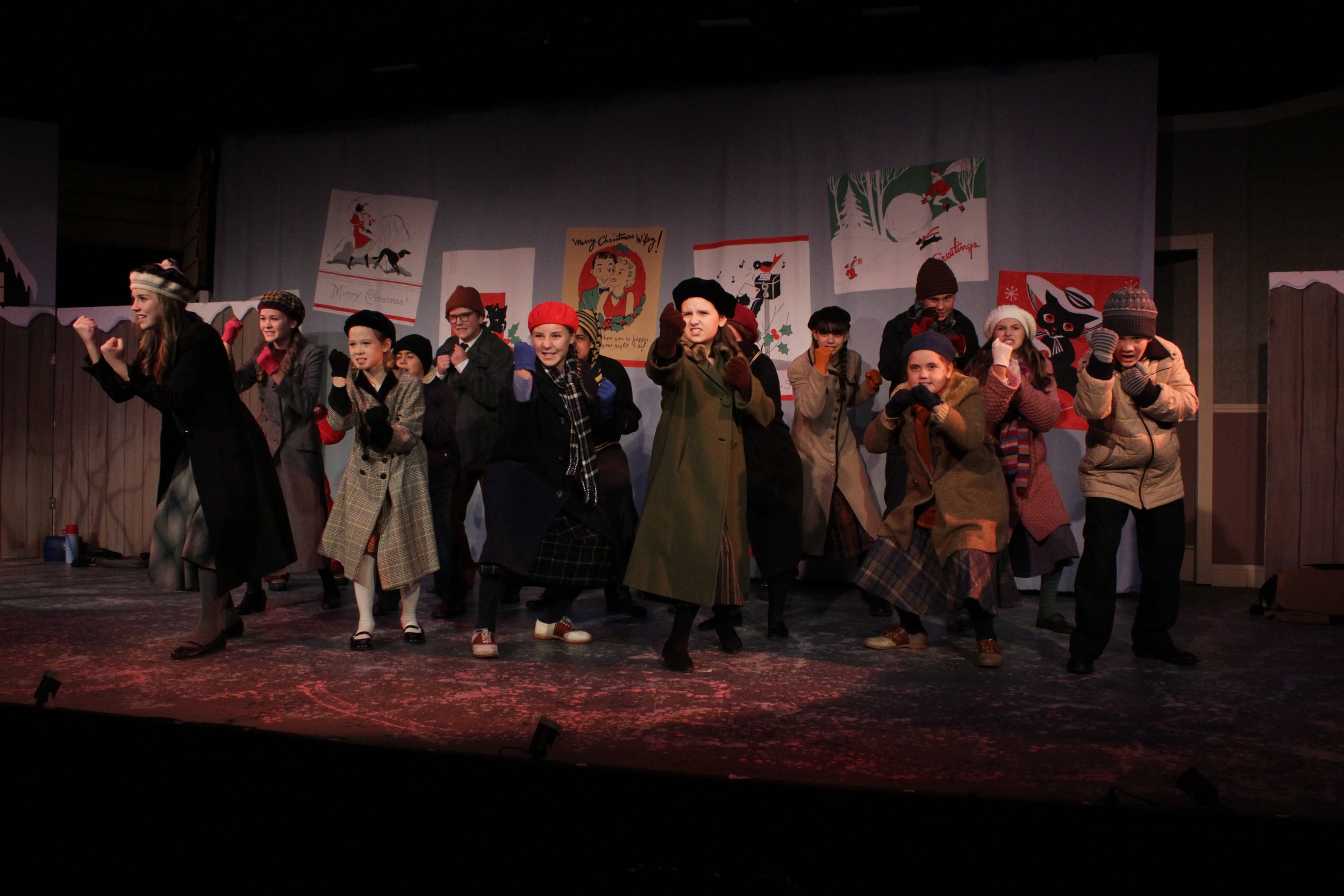 A group of performers on stage dressed in winter clothing, including coats, hats, and scarves, enacting a scene in a play. The background features festive holiday posters.