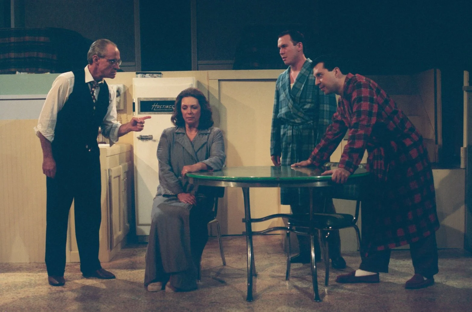 A scene from a theatrical play with four actors. One man, wearing glasses and gesturing, stands beside a seated woman. Two other men, in bathrobes, are standing near a table, engaging with the scene.