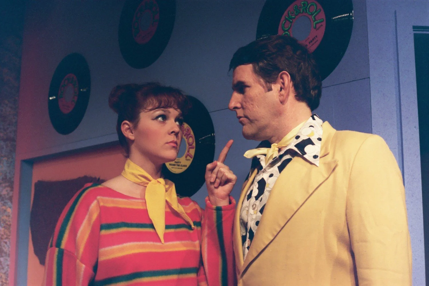 A woman in a colorful striped outfit with a yellow scarf points at a man in a yellow suit. They are standing in front of a wall decorated with vinyl records.