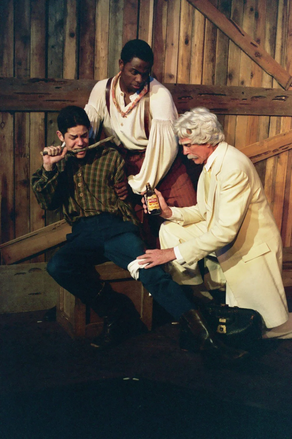 Three actors in theatrical clothing enact a medical scene, with one actor holding a saw to the knee of another, who is seated and biting a tool, while a third actor in white applies a bottle to a bandaged leg. They are set against a rustic wooden bac