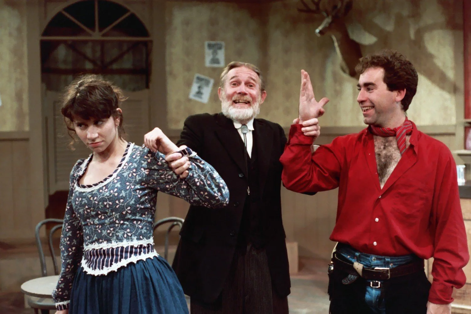 Three actors on stage in a theatrical performance. One woman in a floral dress, a man in a suit, and another man in a red shirt are posing. The background has rustic, vintage decor.