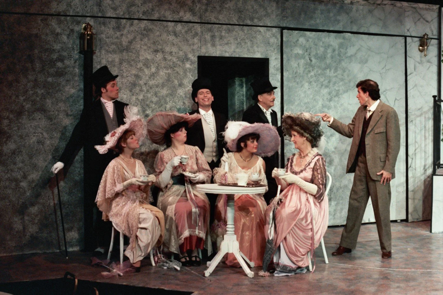 A theatrical performance scene featuring six people dressed in vintage costumes. Three women in elaborate hats and dresses are seated at a table with teacups. Two men in suits and top hats stand behind them. Another man in a suit gestures towards the