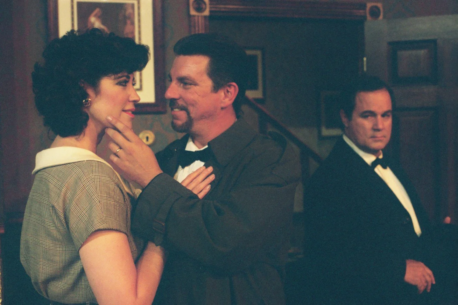 Scene of two men in tuxedos and a woman wearing a collared dress in an indoor setting, with framed art on the wall in the background.
