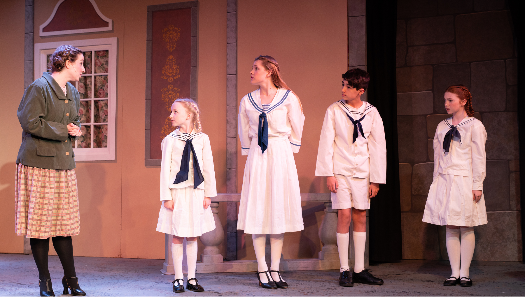 Four children in sailor outfits and a woman in vintage clothing performing a scene on stage in a theatrical production.