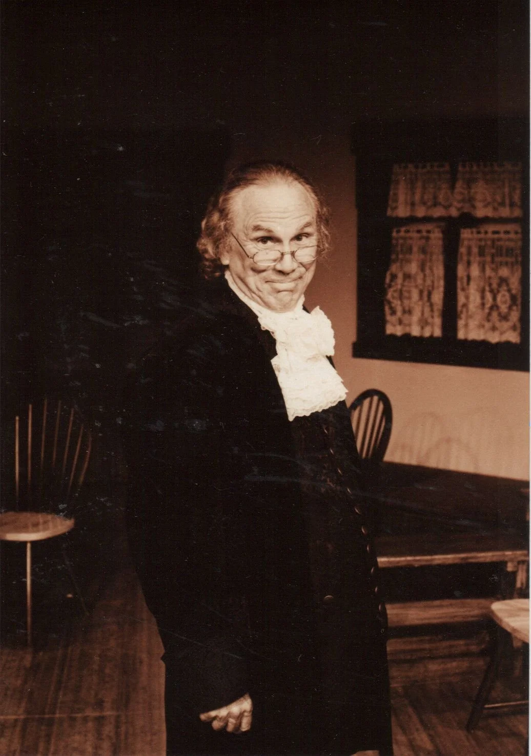 An older man in historical costume with glasses and a white neck ruffle, standing in a room with wooden chairs and a lace-curtained window.