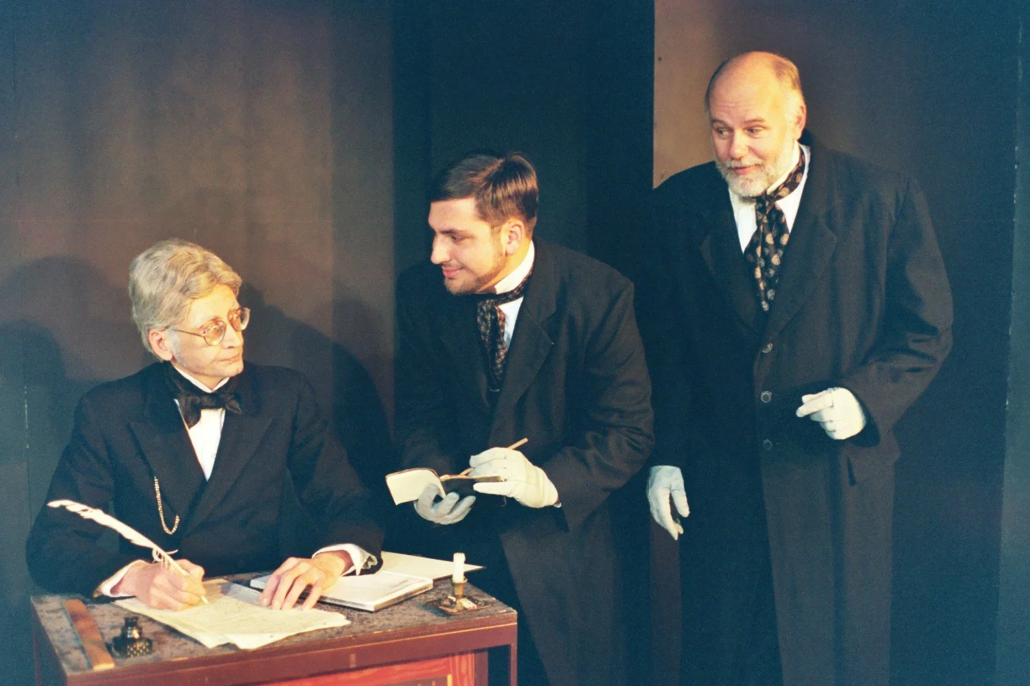 Three men in period costumes and formal attire, with one sitting at a desk holding a quill pen, another holding a notebook, and the third standing nearby, inside a dimly lit room.